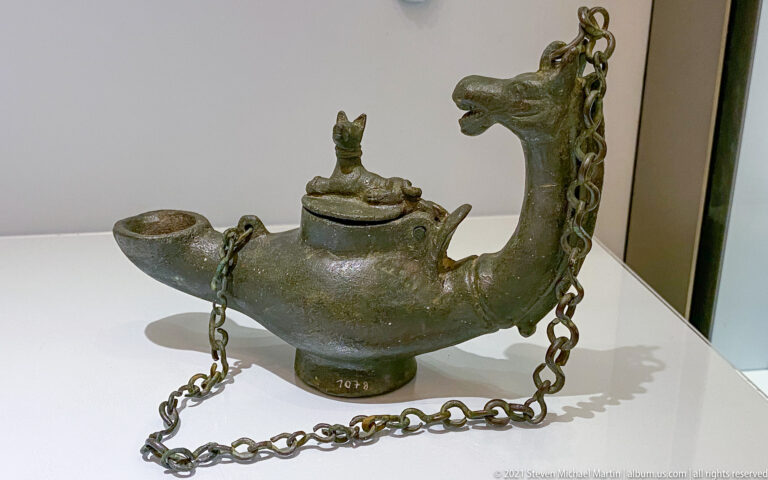 Patinated bronze hanging oil lamp with long nozzle, zoomorphic handle, suspension chains, and small animal on the lid.