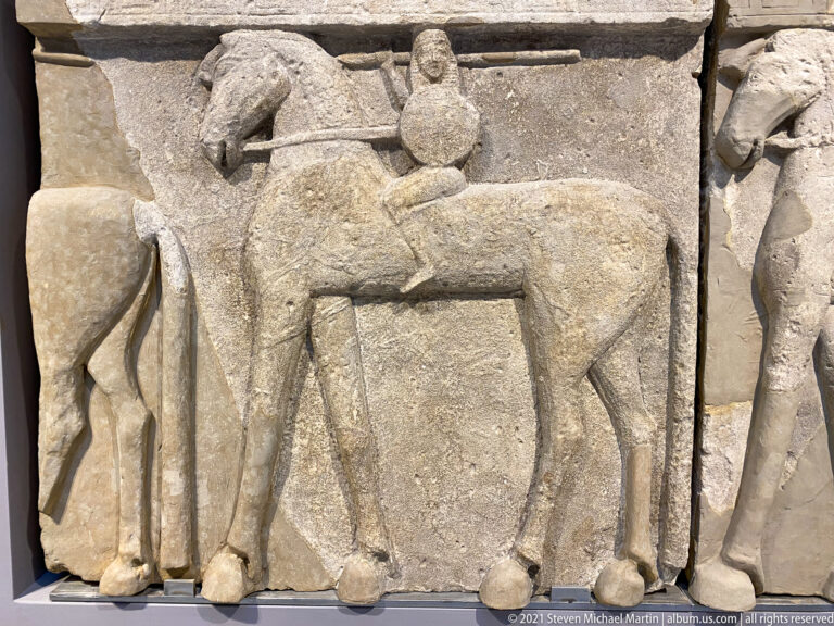 Worn stone relief with a mounted rider on a horse in profile and fragmentary figures at both edges.