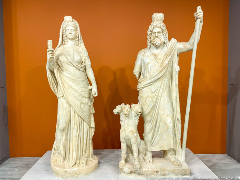 Marble statues of draped Isis-Persephone and bearded Sarapis-Hades with high headdress, staff, and small animal.