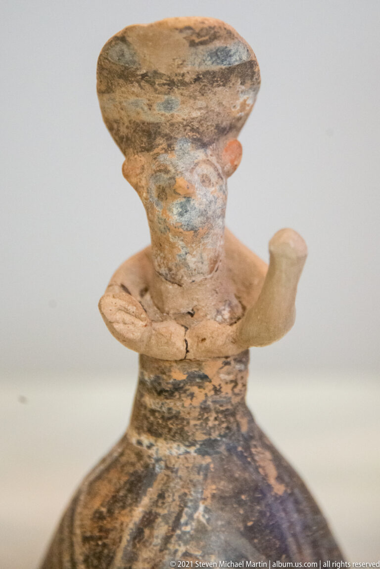 Small ceramic vessel with modeled human head, side arm-like projections, and dark painted bands.