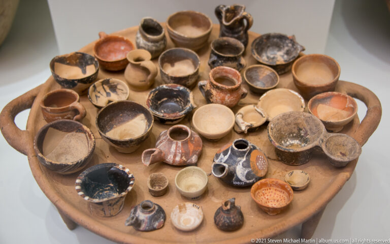 Broad tray filled with dozens of miniature ceramic cups, bowls, jugs, and small handled vessels.