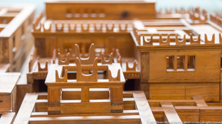 Close detail of a wooden palace model showing upper-story windows, projecting beams, parapet uprights, and adjoining roofs.