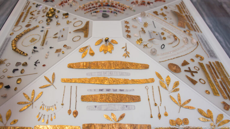 Display case filled with gold strips, leaf-shaped ornaments, beads, pendants, chains, and small fittings.
