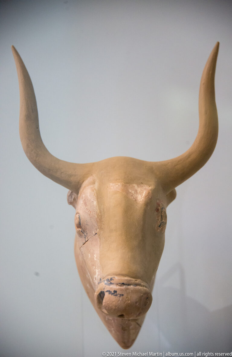 Frontal view of a pale Minoan bull-head vessel with long curved horns, modeled muzzle, eyes, and ears.
