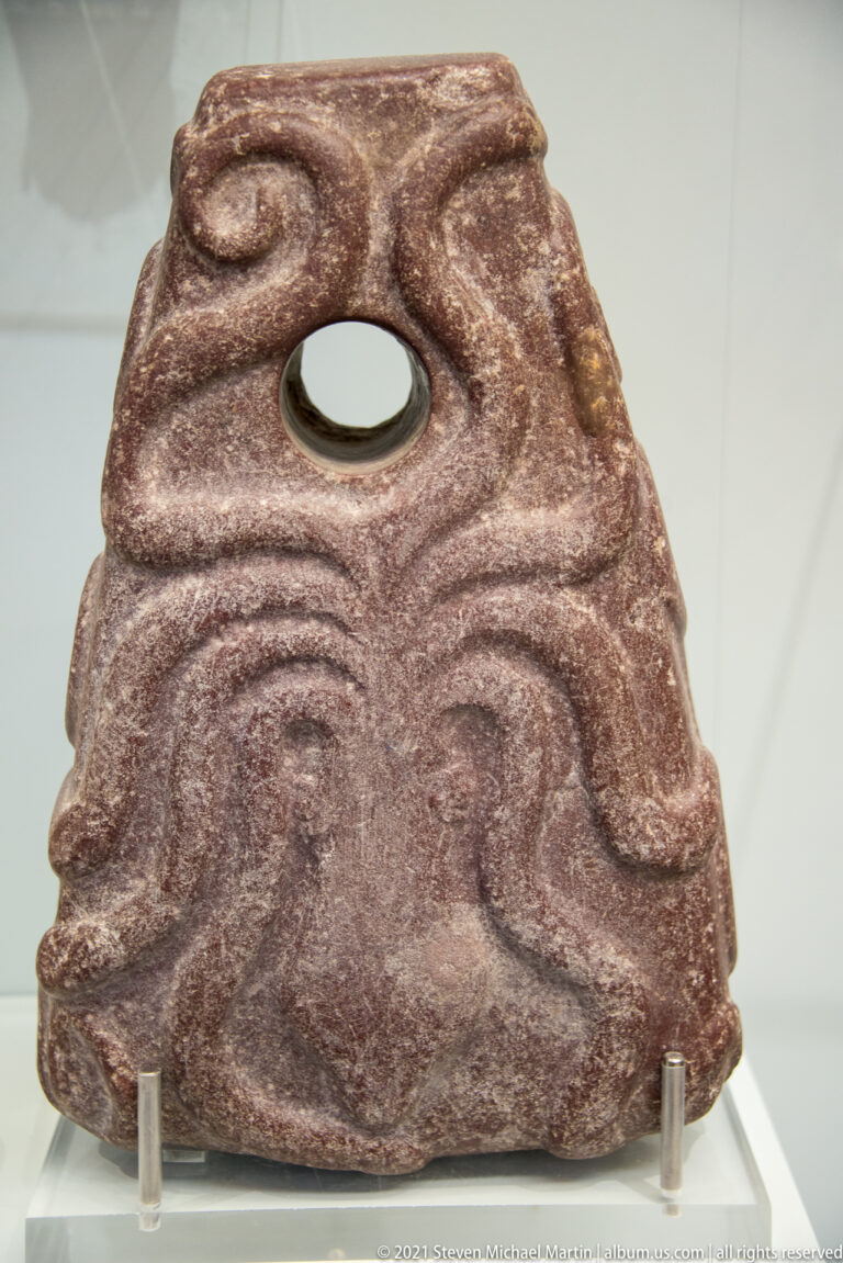 Triangular reddish stone object with a large circular perforation and broad curving relief carving on its face.