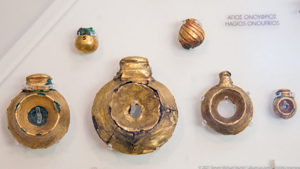 Five small gold-covered circular objects, three with central openings, displayed against a white museum backing.