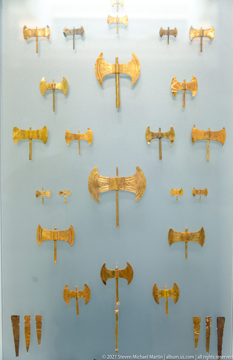 Museum display of numerous small gold double-axe ornaments arranged in rows against a pale background.