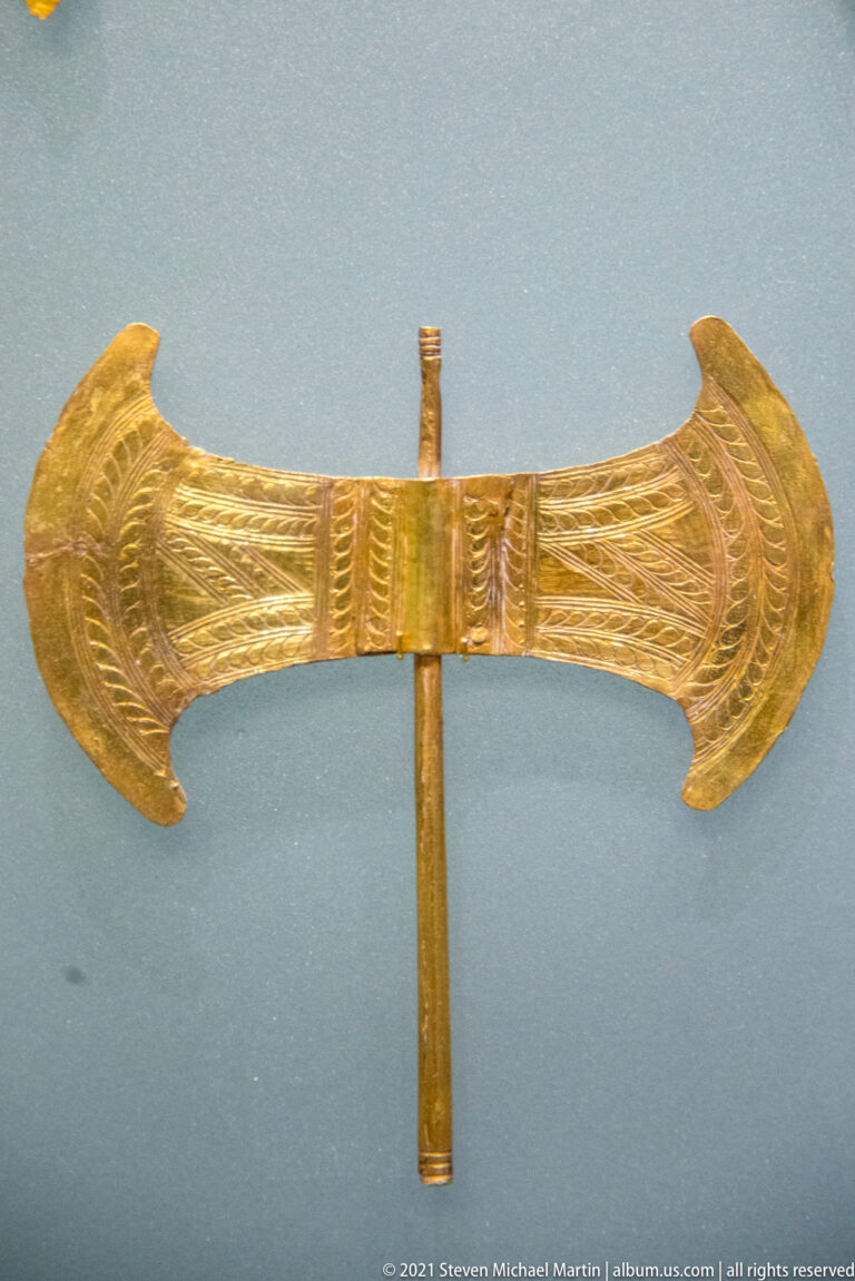 Close view of a gold Minoan double-axe ornament with radiating linear decoration across two symmetrical curved blades.