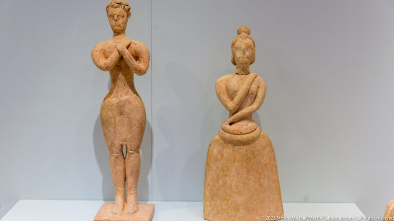 Two Minoan female terracotta figurines, one slender with raised hands and one wearing a long bell-shaped garment.