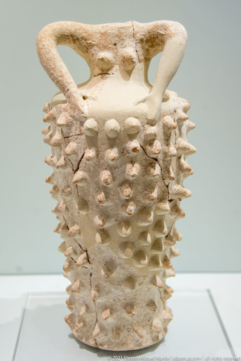 Pale ceramic vessel covered with rows of projecting knobs and spikes, topped by two large loop handles.