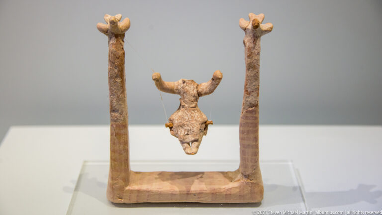 Small terracotta model with two tall upright posts flanking a suspended bull head above a rectangular base.