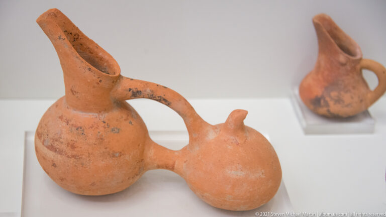 Terracotta vessel with round body, tall spout, large handle, and a smaller joined side chamber.