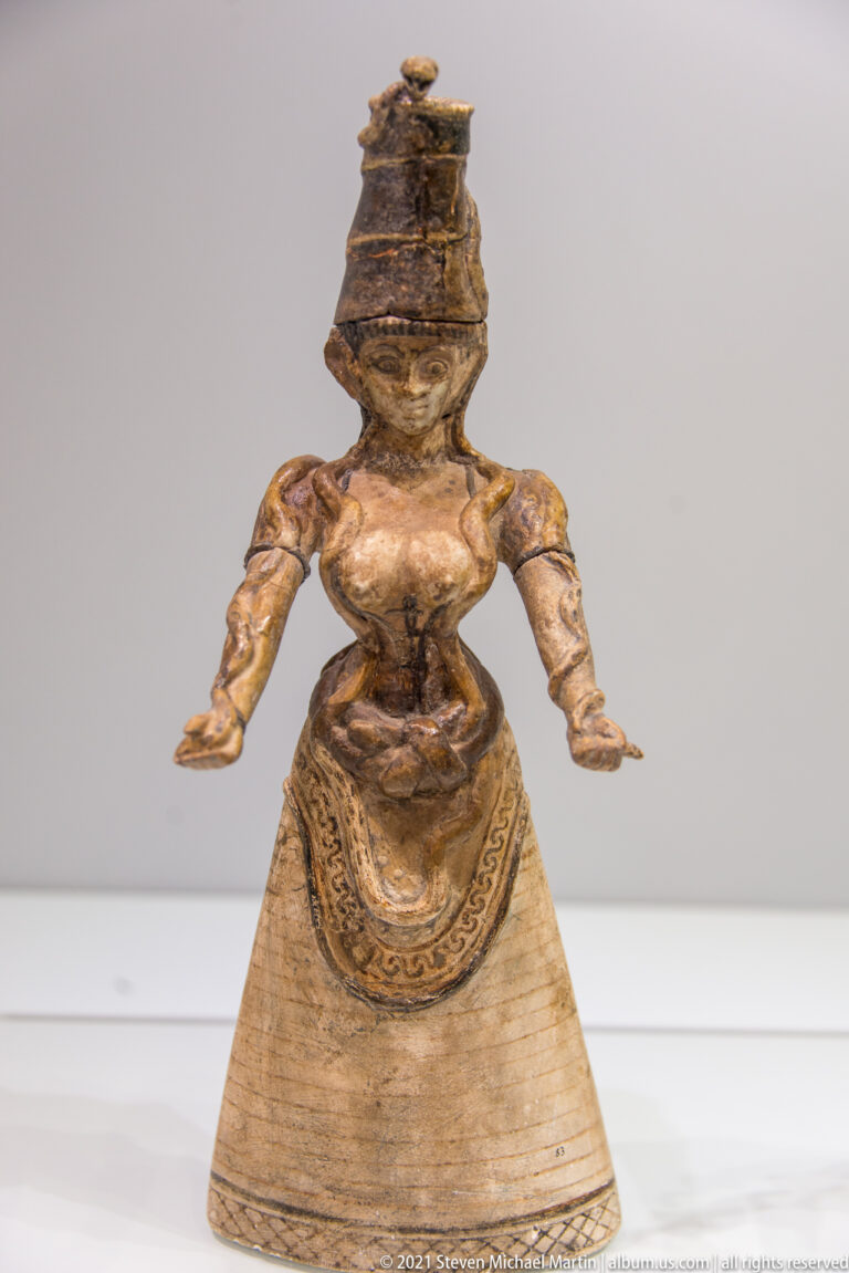 Minoan female figurine with tall headpiece, extended arms, fitted torso, and broad decorated bell-shaped skirt.