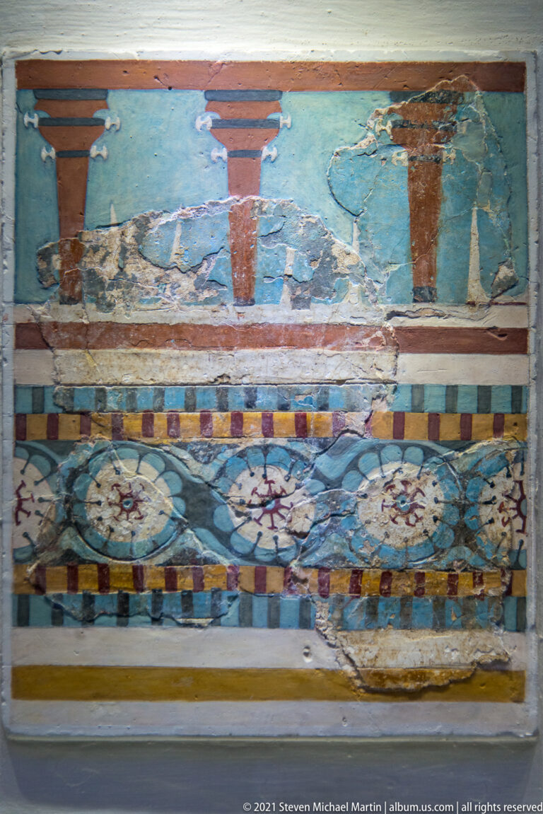 Fragmentary Minoan fresco with blue and red upper register, multicolored borders, and a lower band of large floral rosettes.