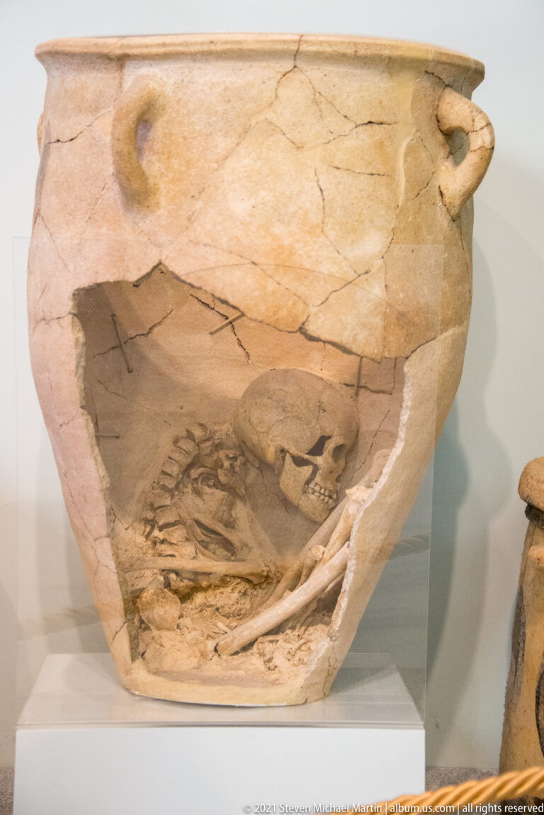 Large cracked ceramic burial jar displayed in cutaway, with human skull and skeletal remains visible inside.