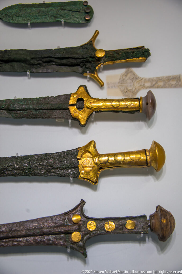 Horizontal museum display of corroded bronze swords and daggers with several ornate gold-covered hilts, guards, and pommels.