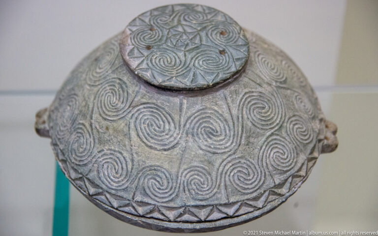 Round dark lidded ceramic vessel covered with incised spirals and triangular border patterns.