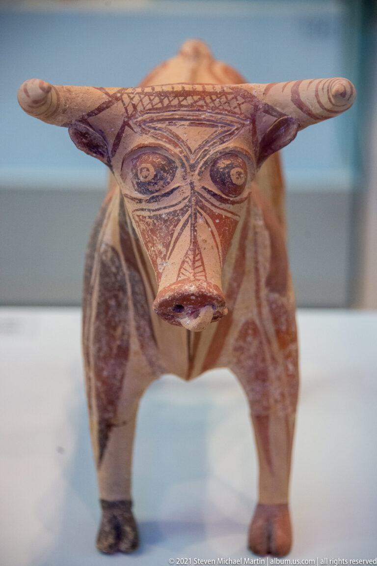 Painted Minoan bull figurine with broad horns, outlined eyes, pointed muzzle, four legs, and geometric body decoration.