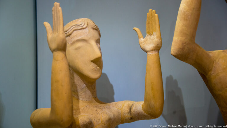 Close view of a terracotta female figurine with both hands raised beside the head and a stylized face.