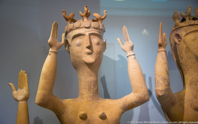 Terracotta female figurine with both arms raised and a tall branching headdress above a banded coiffure.