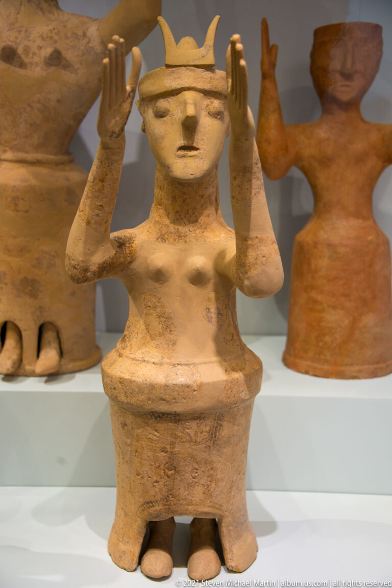 Terracotta female figure with raised hands, angular crown, broad flaring base, and visible feet.