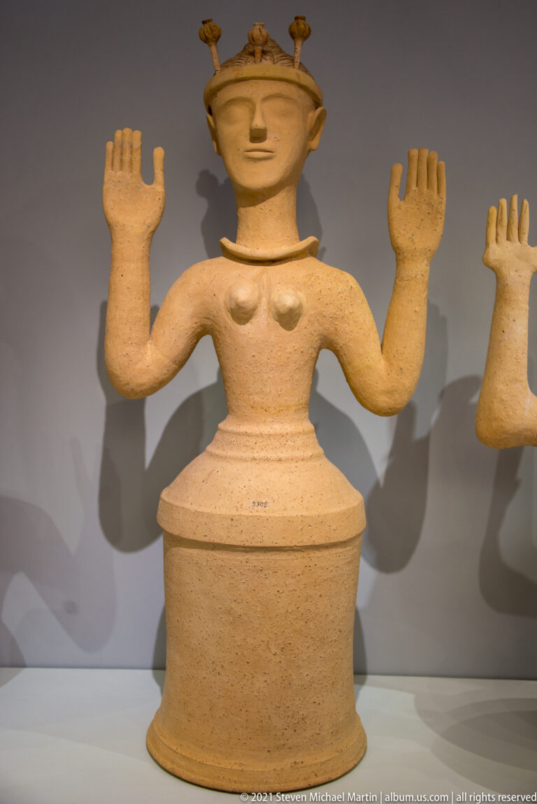 Large terracotta female figure on a cylindrical base with raised palms and a projecting crown.