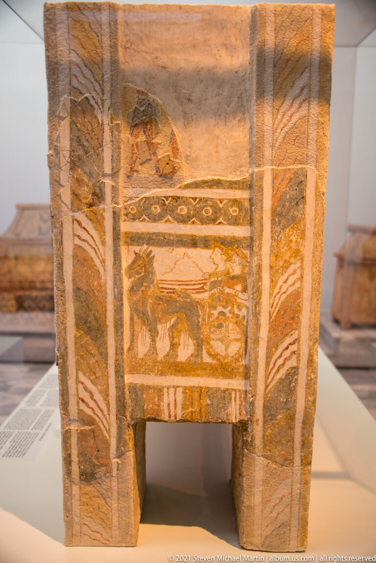 Front view of a painted terracotta larnax on short legs, with framed animal imagery and patterned borders.