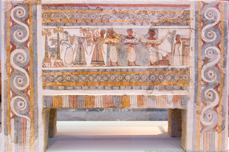 Painted terracotta larnax with a horizontal row of human figures framed by spiral and geometric borders.