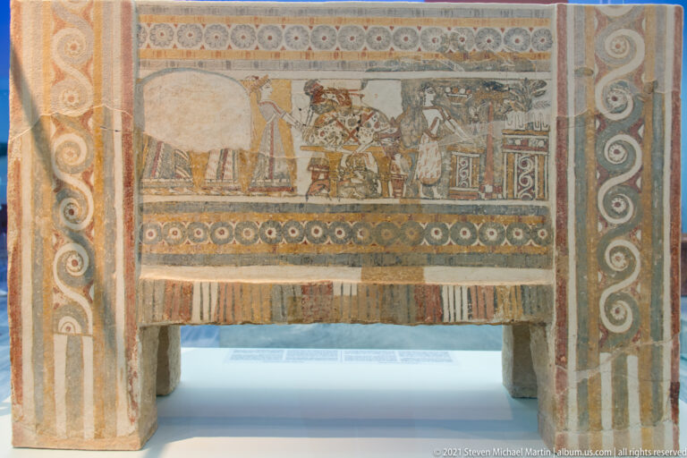 Painted larnax with a multi-figure narrative panel surrounded by rosette, spiral, striped, and geometric borders.
