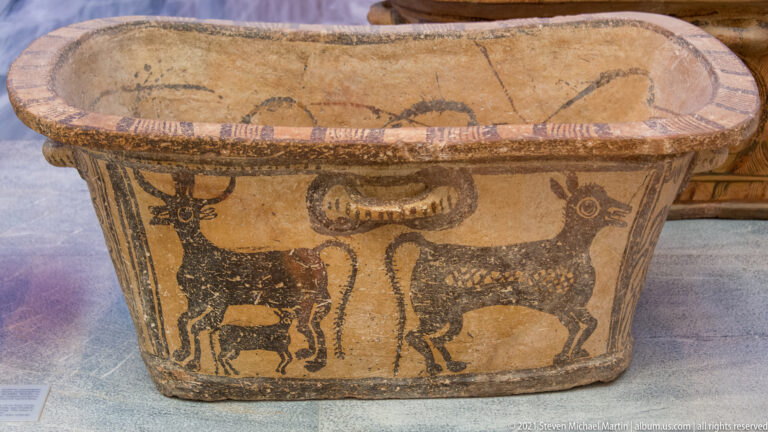 Terracotta larnax painted with two horned bulls facing inward around tall plant motifs on a pale ground.