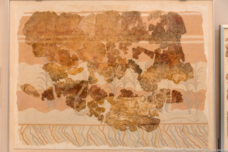 Fragmentary Minoan fresco with reddish and blue-gray plant forms above a wavy painted border.