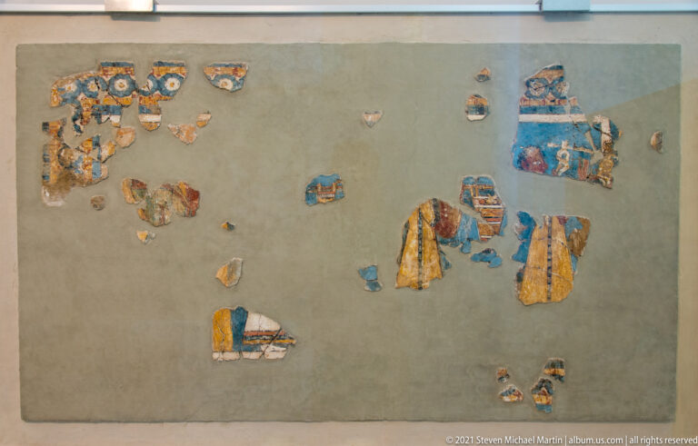 Scattered painted-plaster fragments with blue, yellow, red, black, and white details mounted on a neutral backing.