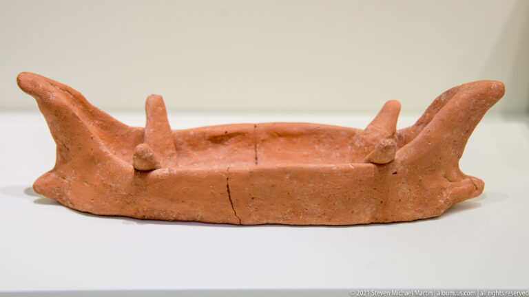 Small terracotta boat model with open hull, high curved ends, and raised projections along the rim.