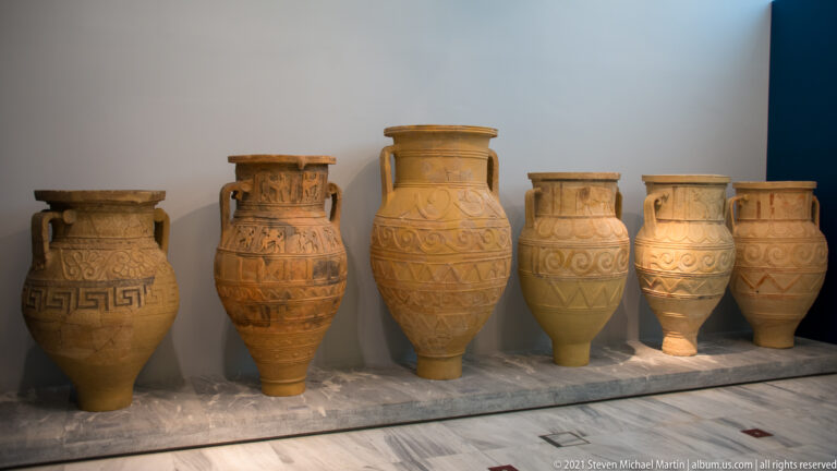 Six tall ceramic storage jars displayed together, decorated with relief spirals, geometric bands, and applied motifs.