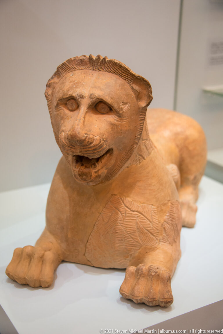 Terracotta lion crouching with forepaws extended, open mouth, broad mane, and incised details across the body.
