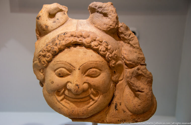 Terracotta face fragment with bulging eyes, broad nose, curling moustache, smiling mouth, and textured hair or headband.