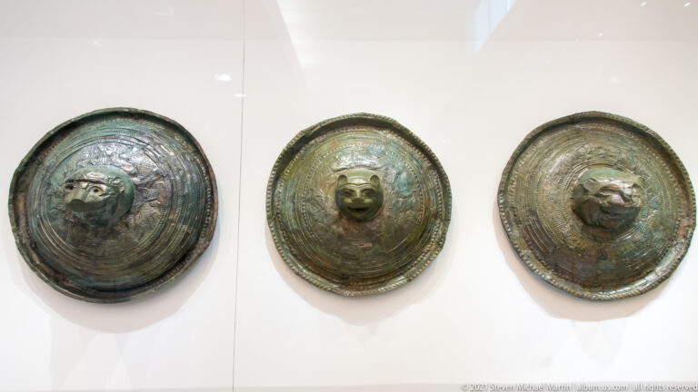 Three patinated circular bronze shields with projecting animal-head bosses and concentric bands of embossed decoration.