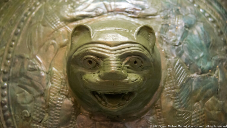 Close view of a patinated bronze shield with a projecting snarling feline head and surrounding embossed figures.