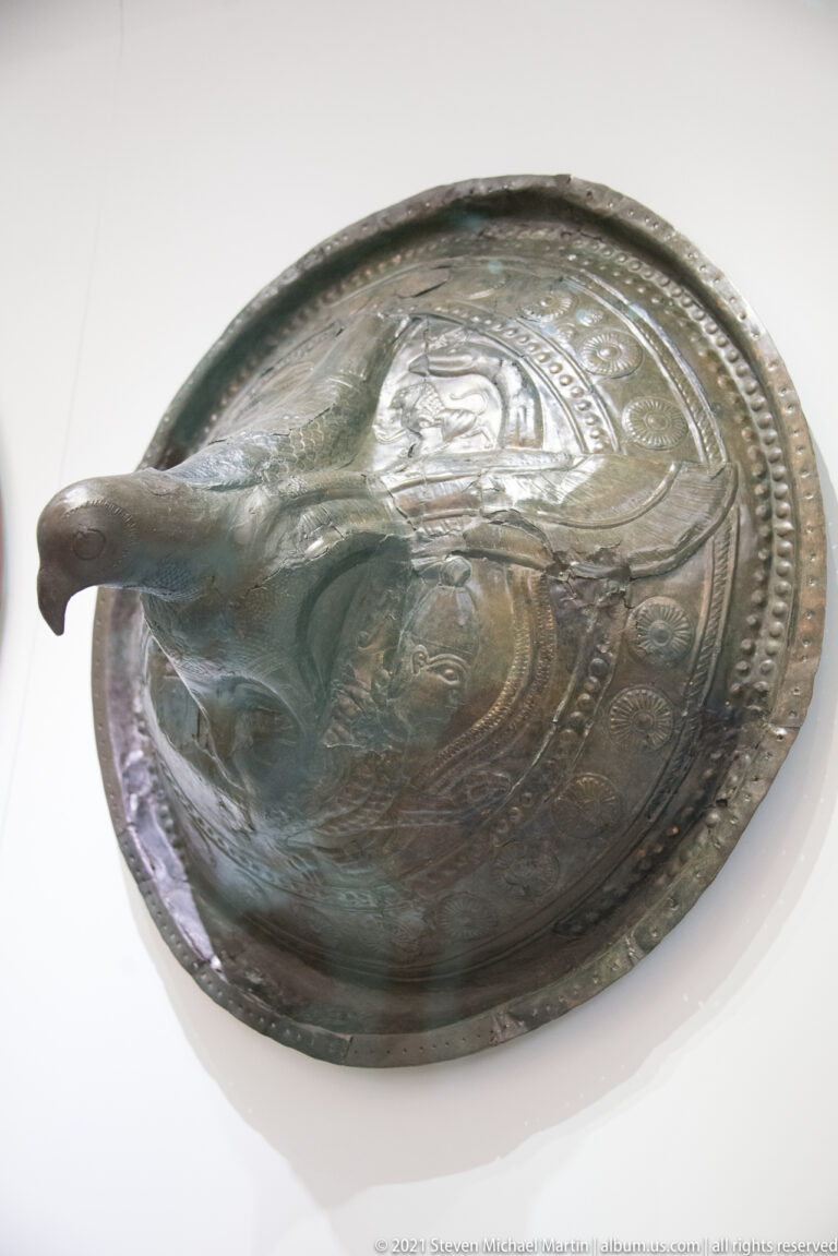 Oblique view of a circular bronze shield with a long projecting central boss and concentric embossed decoration.