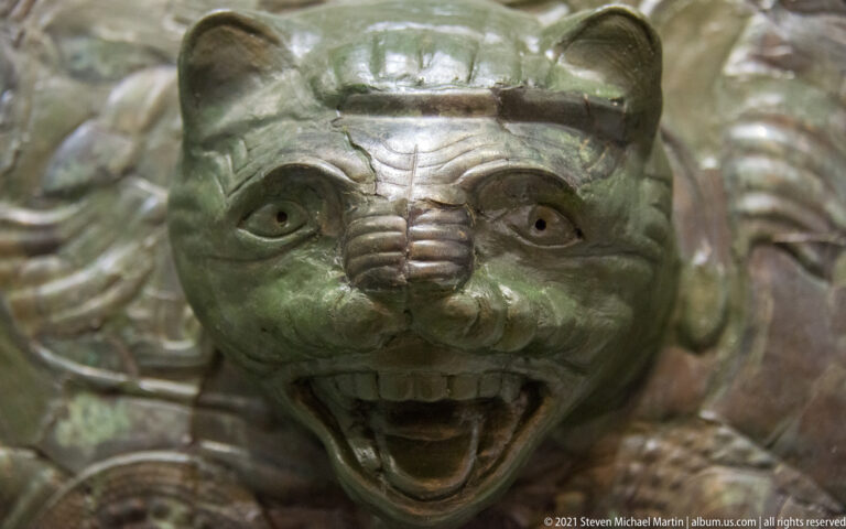 Close-up of a snarling feline head in high relief at the center of a patinated bronze shield.