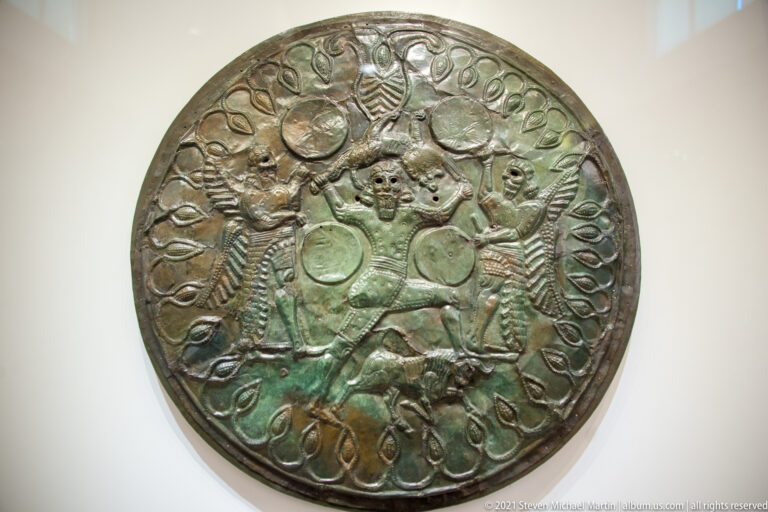 Round patinated bronze shield covered with embossed figures, animals, looping motifs, and a dense central narrative group.