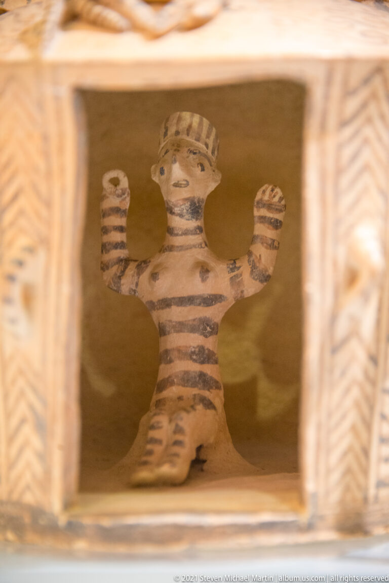 Small striped terracotta human figure with raised arms standing inside the rectangular opening of a painted architectural model.
