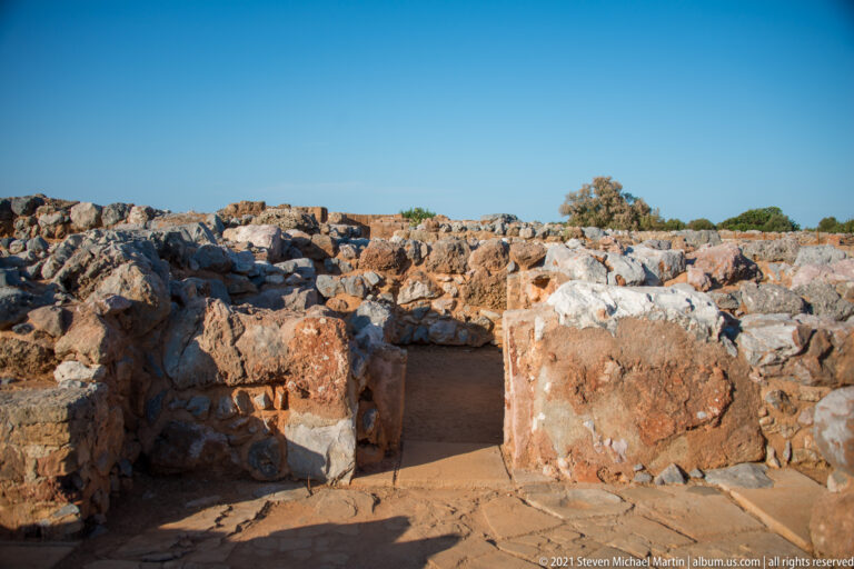 Small excavated space with surviving reddish plaster on low stone walls and an earthen floor.
