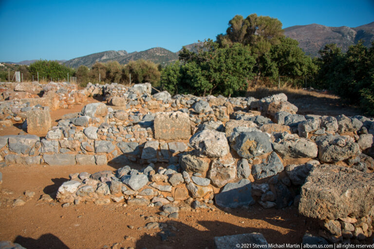 Low palace foundations amid vegetation with a mountain ridge rising in the background.