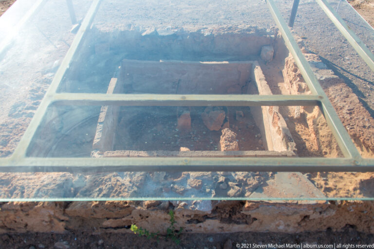 Rectangular excavated feature visible beneath a glass protective cover with a metal frame.