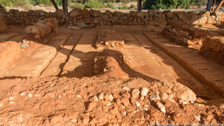 Roofed excavation with an earthen floor, low stone walls, and a narrow raised feature through the center.
