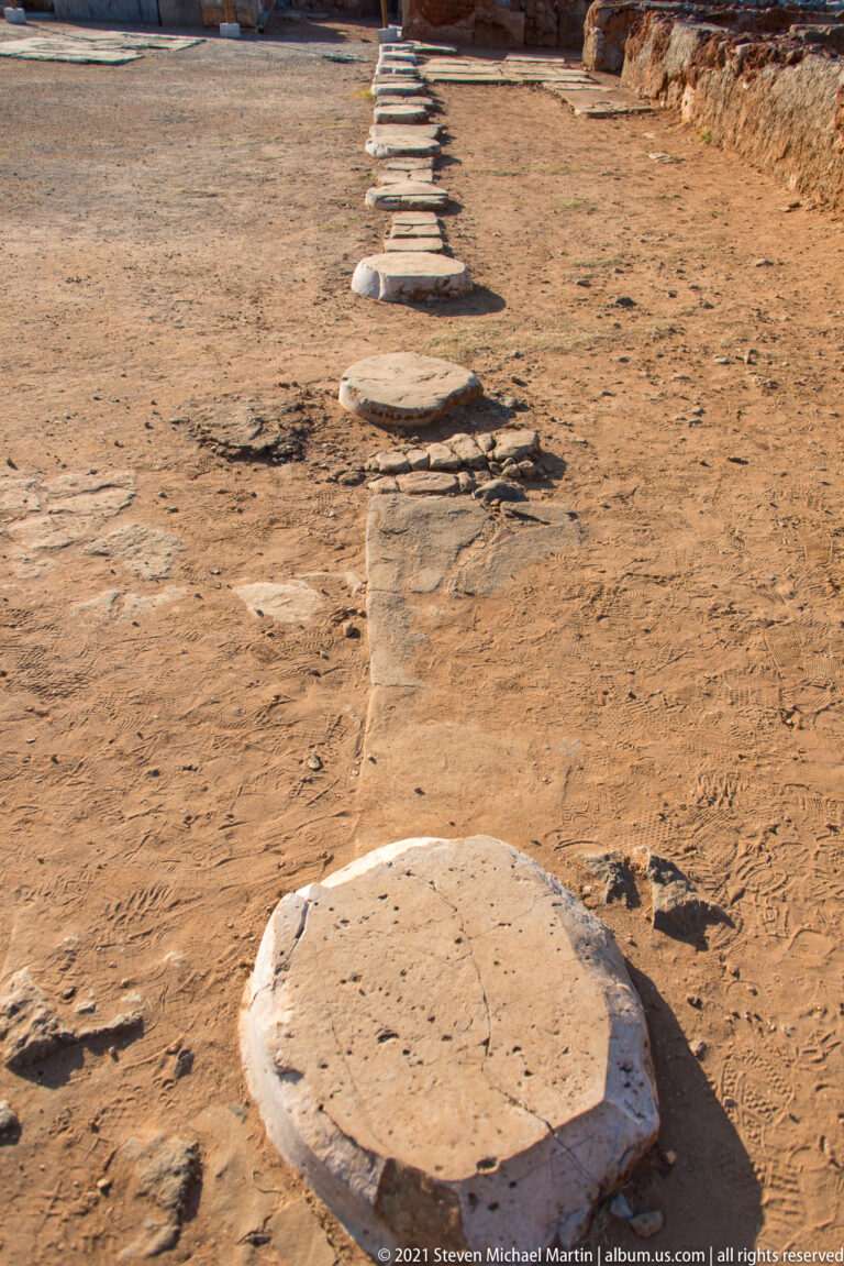 Row of circular stone column bases receding across an open earthen surface at Malia.