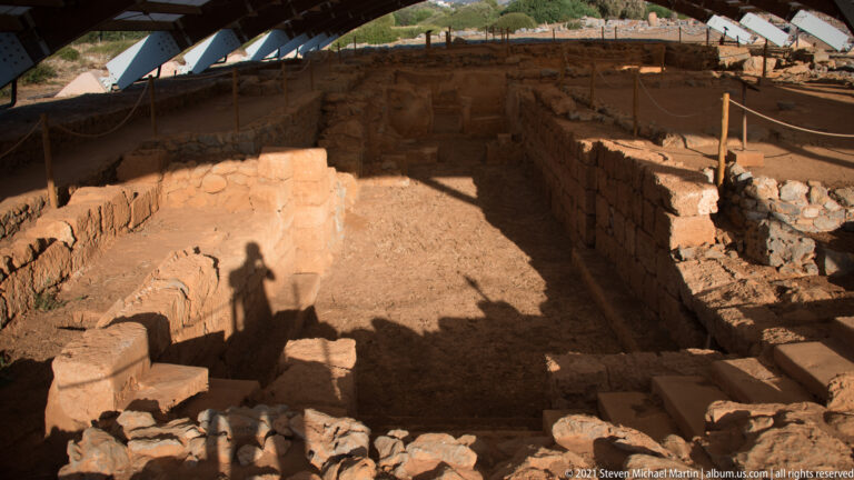 Deep rectangular excavated chamber beneath a shelter, with the photographer’s shadow across the foreground.