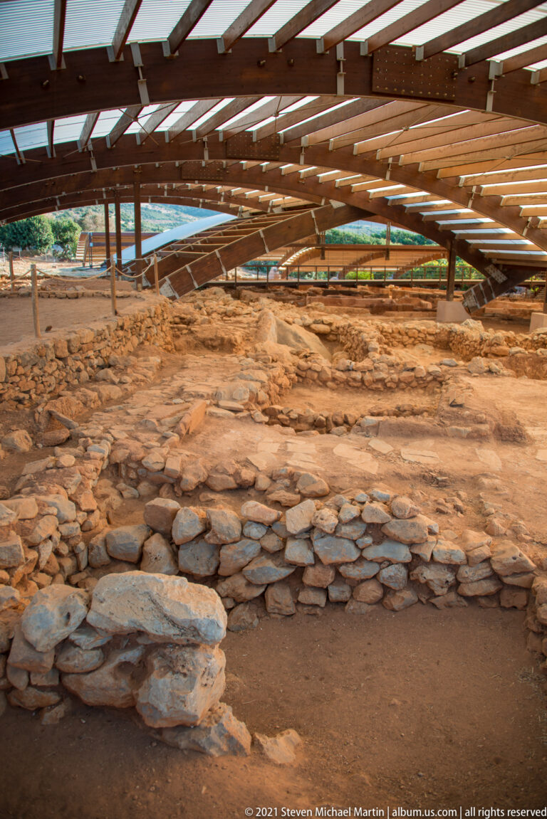 Low curved and straight stone walls forming shallow excavated spaces beneath a timber shelter.