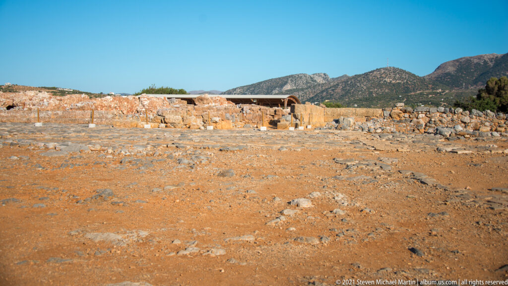 Low stone foundations across the Malia archaeological site with a mountain ridge under a clear blue sky.
